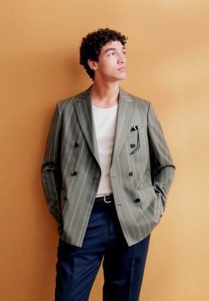 Junger Mann mit lockigem Haar, der einen gestreiften olivgrünen doppelreihigen Blazer, ein weißes Hemd und marineblaue Hose trägt und seitlich gegen eine gelbe Wand blickt.