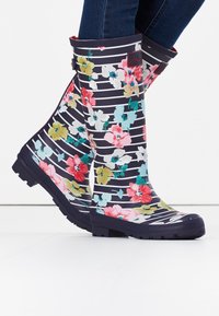 Marine rubberlaarzen met witte strepen en kleurige bloemenprint, gedragen met donkerblauwe jeans op een witte vloer.