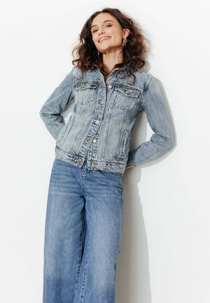 Jeune femme souriante, portant une veste en jean bleu clair et un jean bleu à jambes larges, debout devant un fond blanc, les mains derrière le dos.