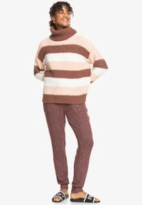 Gruby sweter z dzianiny w różowe i brązowe paski, wysoki kołnierz typu golf, zestawiony z burgundowymi spodniami dresowymi i czarnymi klapkami z białym logo.