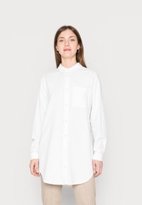 Camisa branca de botões, feita de tecido texturizado, com colarinho, mangas longas, um bolso no peito e uma bainha arredondada.