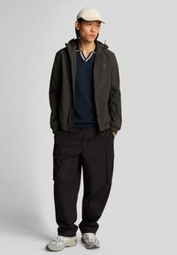 Giacca nera con zip, shirt blu navy, pantaloni cargo neri e sneakers grigie. Cappello chiaro. Tessuto morbido, vestibilità rilassata.