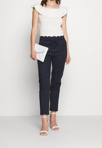 Žena oblečená v bílém bezramenném topu s volánovým výstřihem, tmavě modrých kalhotách s gumičkou, bílých lodičkách na vysokém podpatku a držící bílou kabelku.