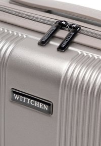Valigia rigida con cerniera in grigio metallizzato con texture a righe, dotata di una placca con logo WITTCHEN nera e doppie linguette nere per la cerniera.
