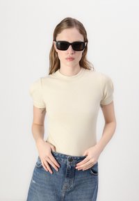 Bež zadnjikast kratki rokav top s visokim ovratnikom, kombiniran z jeans krilom. Model nosi črne pravokotne sončna očala in ima dolge lase.
