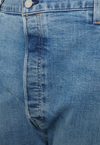 Jeans de ganga azul-claro com uma textura ligeiramente desbotada, apresentando costura laranja e um botão de metal na cintura.
