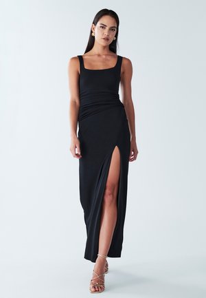 Black maxi dress with a square neckline, ruched design at the waist, and a side slit. Complemented by strappy gold sandals.