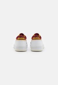 Fehér tornacipők piros és sárga sarokpárnával, sima textúrával és sárga "Reebok" logóval a hátsó részen.