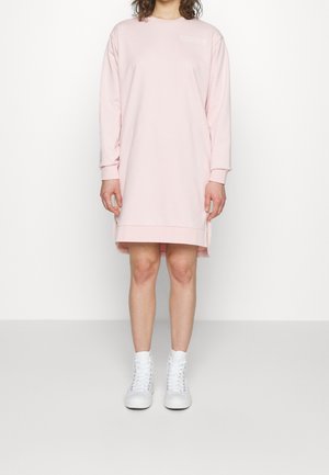 Person trägt ein blassrosa Sweatshirtkleid mit langen Ärmeln und Seitenschlitzen sowie weiße hohe Sneakers und steht vor einem schlichten weißen Hintergrund.