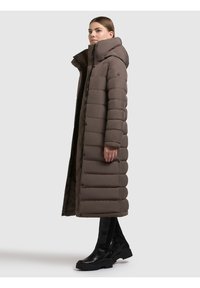 Palton lung, căptușit, de culoare maro închis, cu guler înalt și nasturi. Are un finisaj neted, texturat și este asortat cu ghete negre până la gleznă.