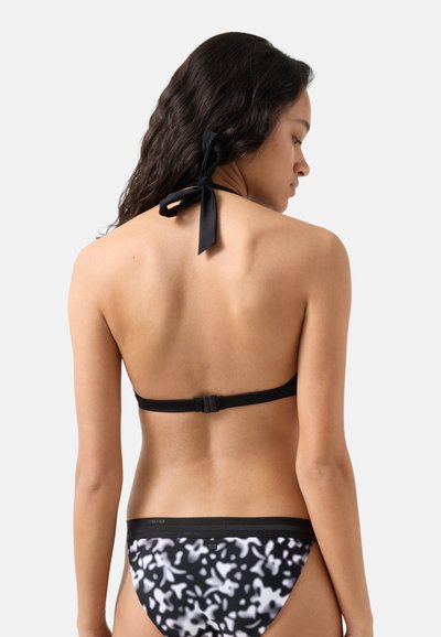 Kvinde med langt mørkt hår iført sort halterbikinitop og sort-hvide mønstrede bikinibund, set bagfra.