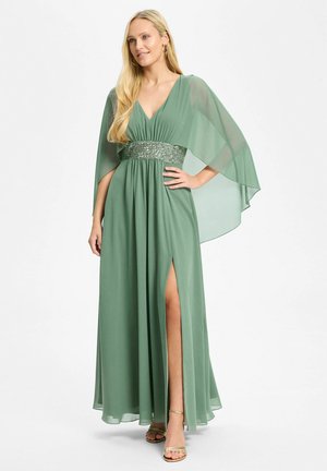 Femme portant une longue robe vert sauge avec des manches cape en voile, une ceinture à sequins et une fente haute à l'avant, associée à des talons dorés à brides.