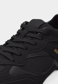 Μαύρο αθλητικό παπούτσι με υφασμένο μπροστινό μέρος με κορδόνια, γλώσσα από διχτυωτό υλικό και λογότυπο "HUGO" σε κίτρινο στην πλαϊνή επιφάνεια.