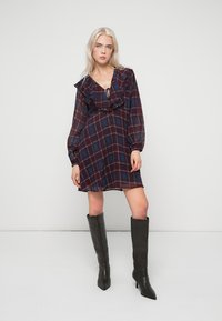 Šaty s dlouhým rukávem v kostkovaném vzoru v tmavě červené a modré barvě s volánkovým výstřihem. Doplněné černými, špičatými kozačkami ke kolenům. Hladká textura.