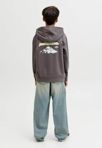 Giovane in piedi con il lato posteriore visibile, indossa una felpa scura con grafica di montagne e jeans blu chiari e larghi.