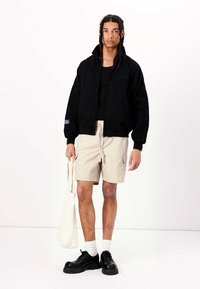 Svart hettejakke med en strukturet stoff, beige shorts, svarte klumpete sko og hvite sokker. Holder en kremfarget totebag.