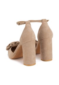 Sandalias de tacón de ante beige con tacones gruesos en bloque, correa en el tobillo y detalle de lazo en la parte delantera. Textura lisa y diseño de punta redondeada.
