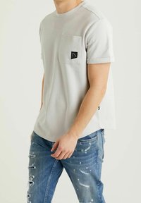 Mężczyzna w jasnoszarej, teksturowanej koszulce z krótkim rękawem i kieszenią na piersi oraz w zniszczonych niebieskich dżinsach z dziurami i plamami farby.