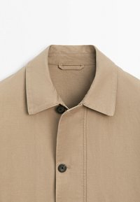 Beige jakke med krave og to synlige sorte knapper, lavet af robust stof, vist fladt på en hvid baggrund.