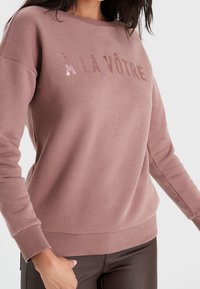 Lyserød sweatshirt med skinnende "À LA VÔTRE" tekst. Har rund halsudskæring, ribstrikkede manchetter og kant samt et blødt bomuldsblanding materiale.