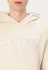 Sudadera beige de tela suave con un logotipo bordado de "GUESS" y "EST. 1981" en un color claro contrastante.