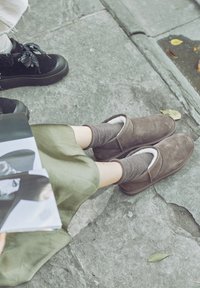 Braune Wildlederhausschuhe mit weißem Innenfutter, getragen mit gestreiften grauen Socken, auf einer Steinoberfläche. Eine teilweise sichtbare Zeitschrift neben den Füßen.