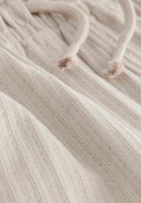Tissu tricoté beige clair montrant une texture subtile avec un détail de cordon de serrage. La surface a une apparence douce et texturée avec des lignes verticales.