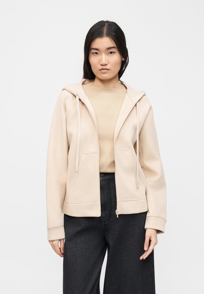 Νεαρή γυναίκα με ίσια μαύρα μαλλιά που φοράει μπεζ φούτερ με φερμουάρ πάνω από μπεζ τοπ και χαλαρά μαύρα παντελόνια, στέκεται μπροστά σε λευκό φόντο.