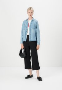 Camisa de denim azul clara com dois bolsos frontais, t-shirt branca com texto vermelho, calças largas pretas, segurando uma mala preta, sapatos pretos.