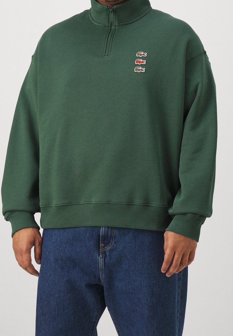 Homme portant un sweat-shirt vert foncé à fermeture éclair quart, avec trois petits logos crocodile et un jean bleu, debout avec les mains détendues.