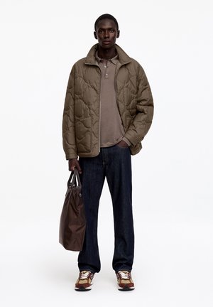 Hombre con chaqueta acolchada marrón, camiseta polo beige, vaqueros oscuros, zapatillas multicolor, sosteniendo un bolso tote marrón oscuro con una mano en el bolsillo.