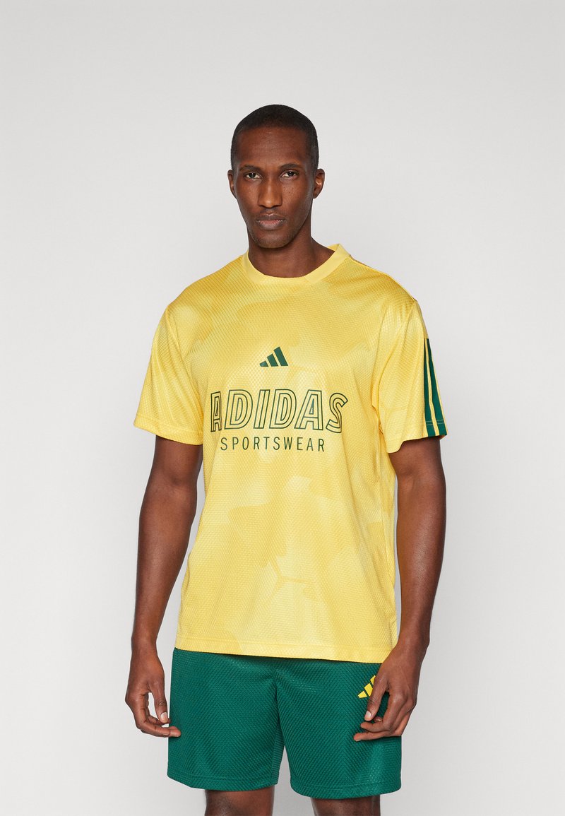 Κίτρινη αθλητική μπλούζα από διχτυωτό υλικό με πράσινο λογότυπο "ADIDAS SPORTSWEAR", συνδυασμένη με σκούρες πράσινες βερμούδες που διαθέτουν ξεχωριστό λογότυπο.