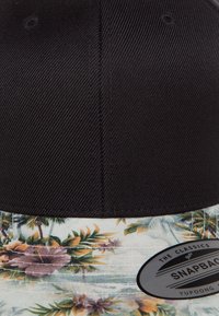 Crna snapback kapa s cvjetnim uzorkom na obodu i potplatu. Oznaka prikazuje "The Classics Yupoong" u kružnom logotipu.