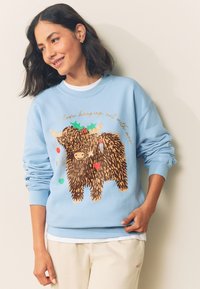 Jasnoniebieska bluza z długimi rękawami, z brązowym obrazkiem futrzanego zwierzęcia i ozdobami świątecznymi oraz złotym napisem nad nimi.