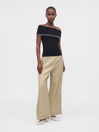 Femme portant un top noir épaules dénudées avec bordure blanche, un pantalon beige large brodé, et des escarpins crème pointus sur un fond uni.