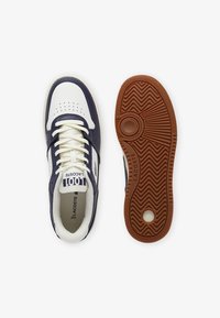 Zapatillas Lacoste en cuero blanco y azul marina, con puntera perforada, suela de goma contrastante y un logo prominente en la lengüeta.