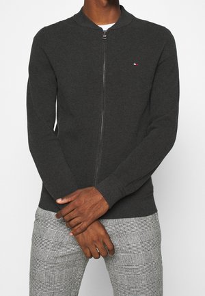 Mann trägt dunkelgrauen Reißverschluss-Pullover und graue karierte Hose, steht mit verschränkten Händen vor weißem Hintergrund.