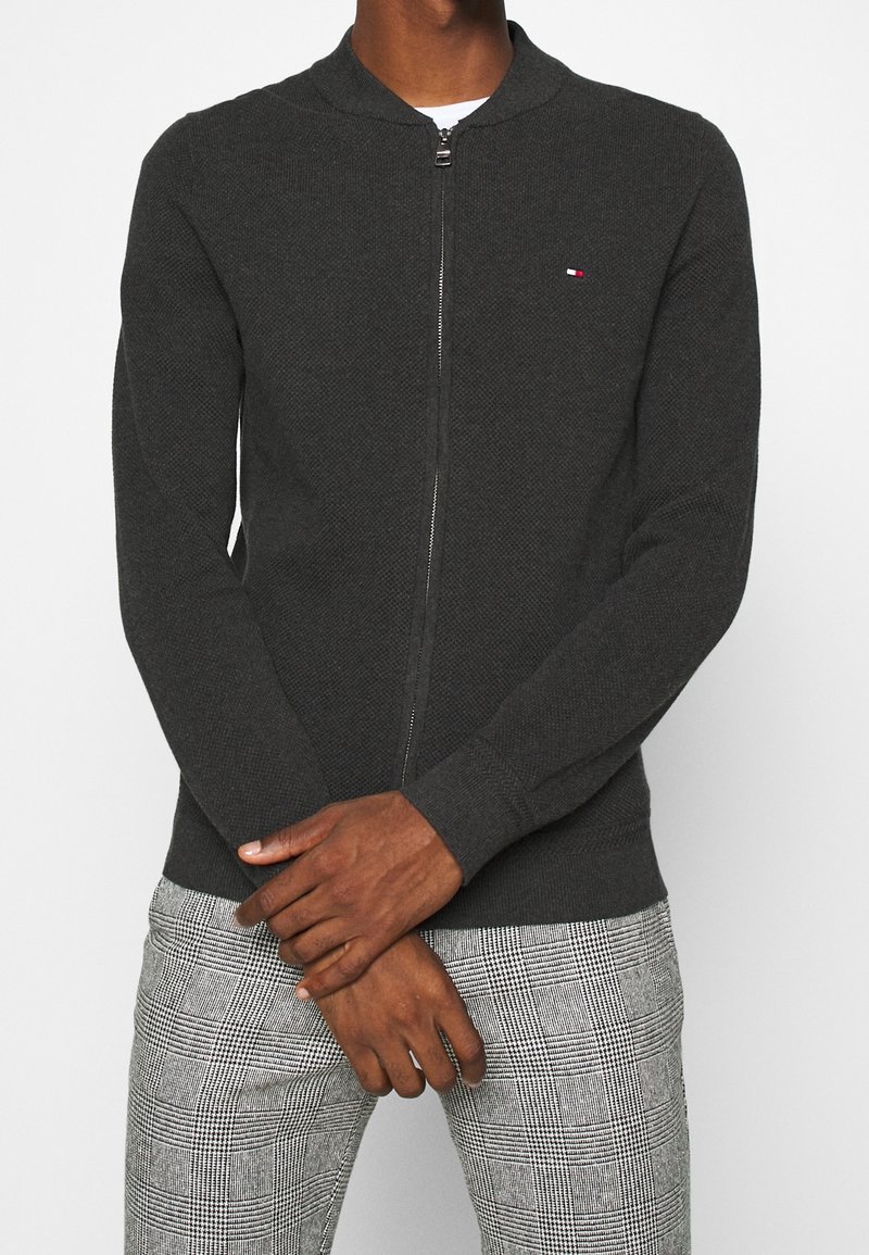 Homme portant un pull à fermeture éclair gris foncé et un pantalon gris à carreaux, debout les bras croisés devant un fond blanc.