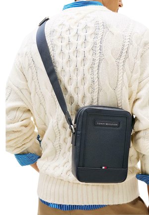 Bolso bandolera de cuero negro granuloso, forma rectangular con cremallera superior, con placa de logo y detalle tricolor en la base.