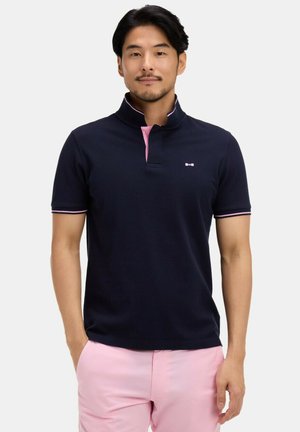 Homme portant un polo bleu marine avec des bordures roses et un short rose clair, debout les mains dans les poches devant un fond uni.