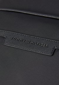 Black fabric backpack featuring a textured black label with embossed "TOMMY HILFIGER" branding. Smooth material with no visible hardware.