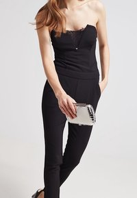 Mono negro sin tirantes con detalles de encaje, silueta ajustada y piernas ajustadas. Sujetando un clutch plateado texturizado con líneas limpias y un acento metálico.