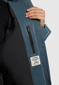 Padded jacket z niebieskim zewnętrzem i czarnym podbiciem, z boczną kieszenią na zamek i metką z tekstem.