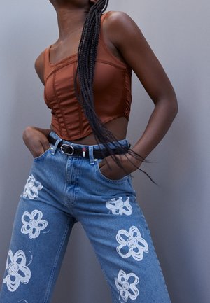 Kvinde med lange fletninger iført en brun ærmeløs korset top og blå jeans med hvide blomstertryk, hænderne i lommerne mod en grå baggrund.