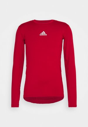 T-shirt de sport rouge à manches longues en tissu stretch, avec un col rond et un logo Adidas blanc sur le devant supérieur. Texture lisse.