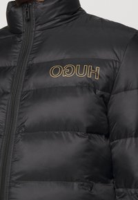 Giacca a vento nera con collo alto, design trapuntato, con logo dorato "HUGO" sul petto e chiusura con zip frontale.