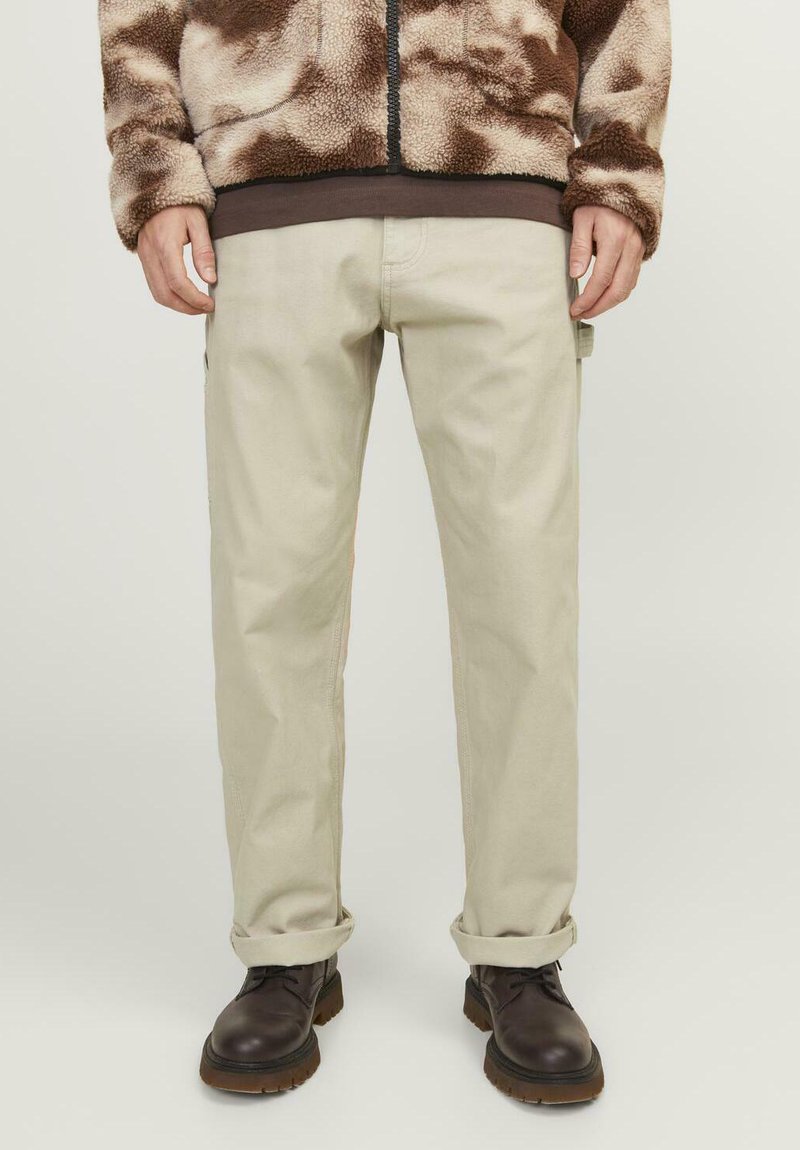 Pantalon beige à coupe droite, avec poches latérales et ourlets retroussés, associé à une veste en polaire marron à motif tacheté. Bottes marron.