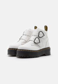 Botas de tobillo de cuero blanco con dos correas ajustables, hebillas en forma de corazón negro y una suela gruesa negra con costuras amarillas.