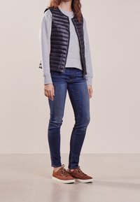 Navy puffer vest over a light gray sweatshirt, paired with dark blue jeans and brown suede sneakers with white soles.
