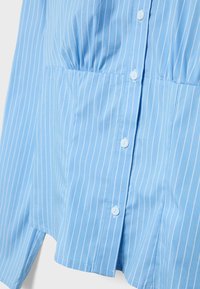 Chemise bleu clair à fines rayures blanches verticales, fermeture à boutons sur le devant, et détail de fronces au niveau de la taille.